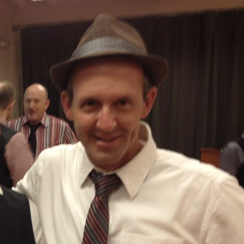 Brett, an Orpheus Male Chorus member, smiling in a fedora hat, tie, and white shirt at a gathering.