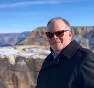 Mike Peterson wearing sunglasses and a dark coat, smiling outdoors with canyon cliffs in the background.