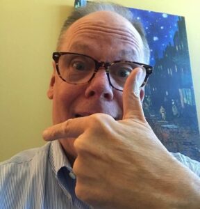 Mike Peterson smiling and pointing at himself while wearing glasses.