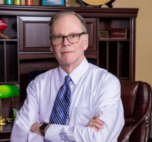 Mike Peterson in a professional portrait, wearing glasses, a white shirt, and a striped tie, seated in an office setting.