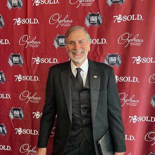 Gary Hapip, an Orpheus Male Chorus member, smiling in a tuxedo in front of a red Orpheus-branded event backdrop.