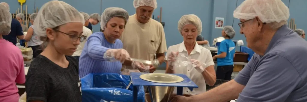 Volunteers of all ages wearing hairnets work together to package meals at a Feed My Starving Children charity event.