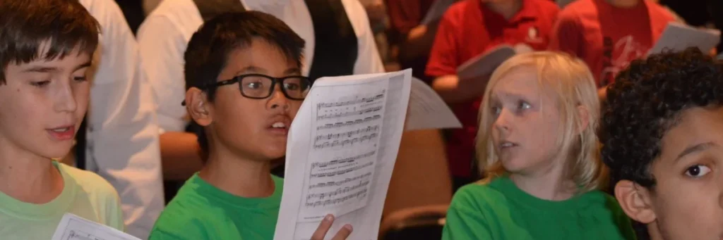 Young choir members in green shirts rehearse with sheet music during a community performance.