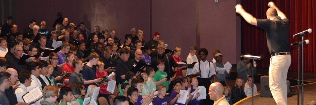 Choir of children and adults rehearsing on stage under the direction of a conductor raising his arms.