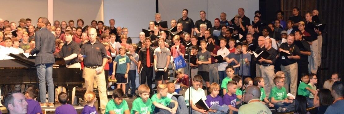 Large intergenerational choir rehearsal with children, teens, and adults performing together on stage.