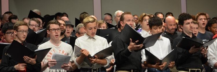 Choir members of different ages singing with sheet music in hand.