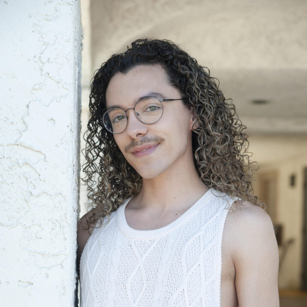 Jacob, an Orpheus chorus member, smiling in a white knit tank top with curly hair and glasses.
