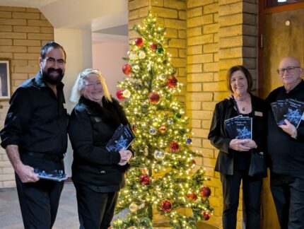 Four volunteers standing by a decorated Christmas tree, holding programs and smiling.
