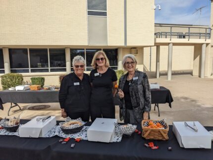 Three volunteers smiling behind a table with food and baskets at an outdoor event.