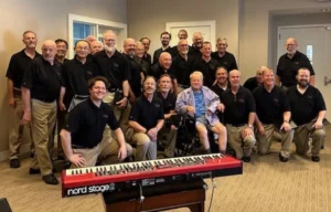 Wayne Grove seated in a wheelchair surrounded by choir members in black shirts, with a keyboard in front.