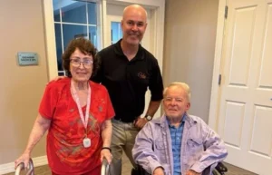 Wayne Grove seated in a wheelchair beside a woman with a walker and a man standing behind them.