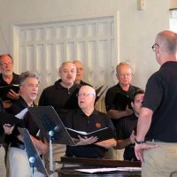 Bob Meier singing with the choir during rehearsal, directed by a conductor.